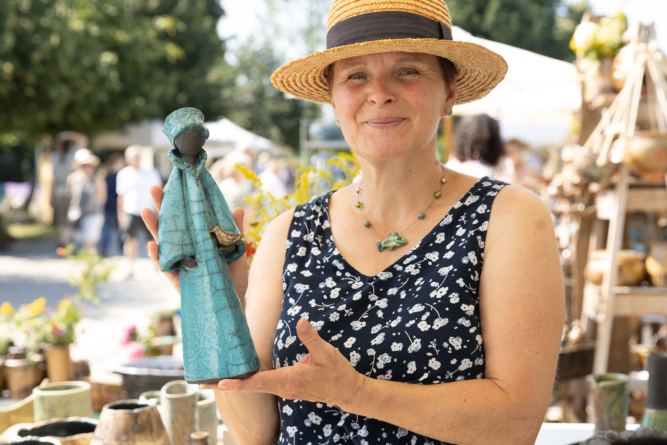 Töpferin mit Strohhut hält eine Keramikfigur mit blauem Gewand und dunklem Gesicht in den Händen, im Hintergrund: Töpferkunstmarkt