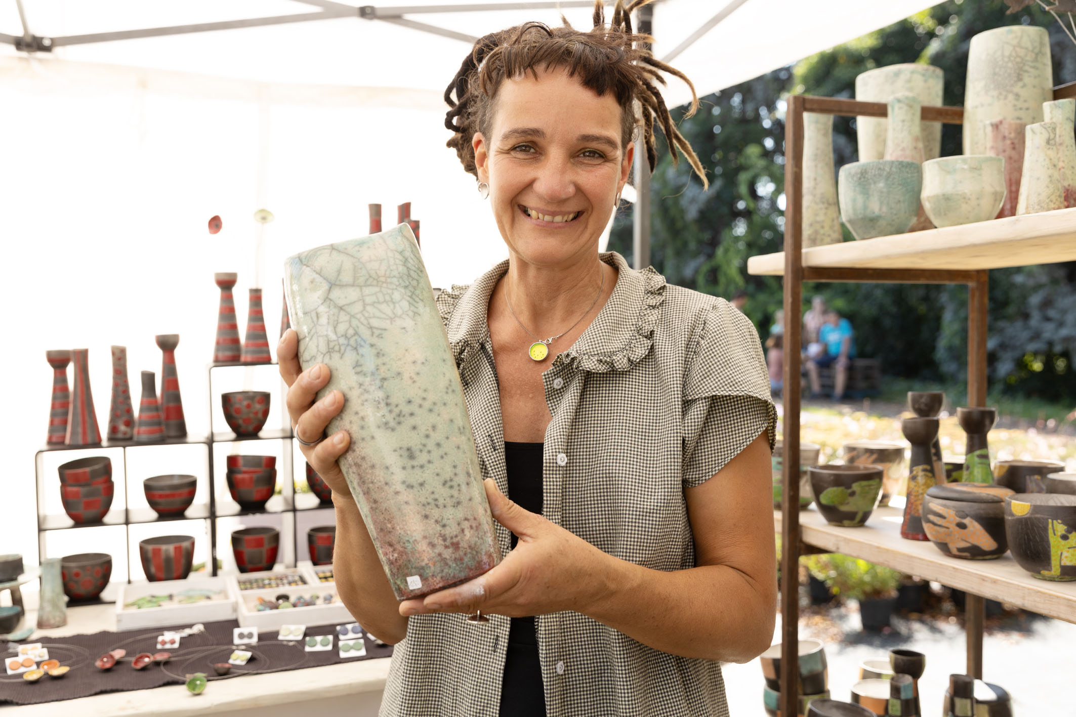 Töpferin mit großer heller Vase in den Händen, lacht in die Kamera, im Hintergrund: weitere Töpferkunst