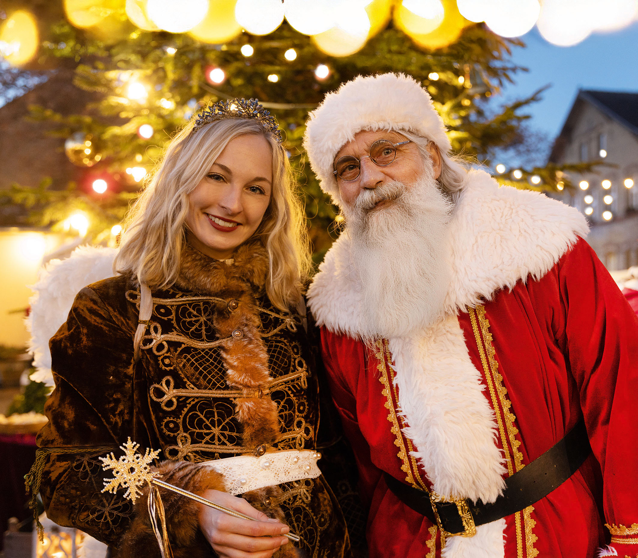 Blonder Weihnachtsengel im barocken Kostüm und Weihnachtsmann mit weißem Bart vor geschmücktem Tannenbaum